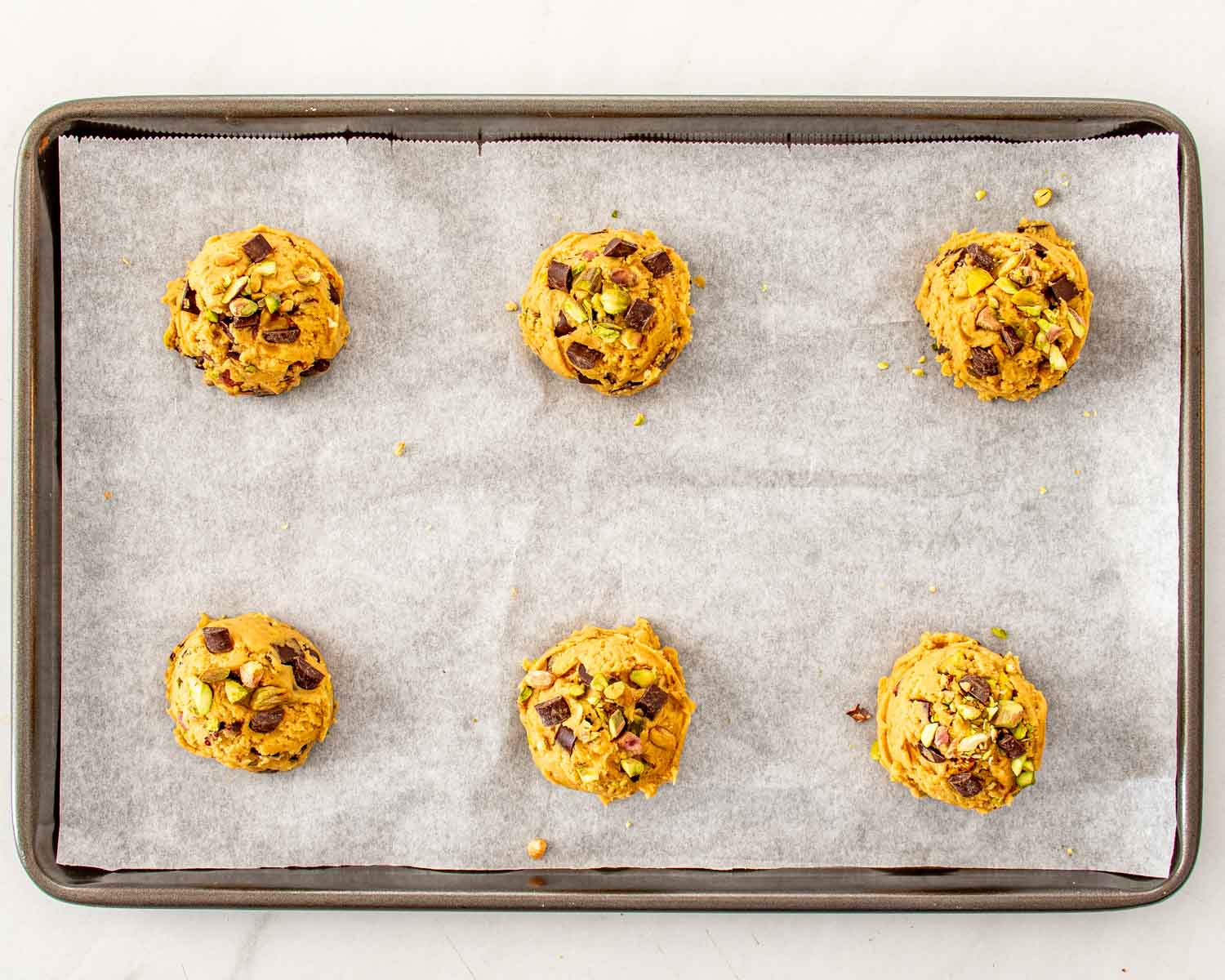 Six chilled pistachio chocolate chunk cookie dough mounds spaced widely apart on a parchment-lined baking sheet before baking.