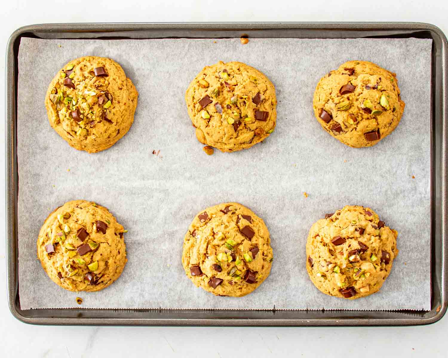 Six baked pistachio chocolate chunk cookies arranged evenly on a parchment-lined metal baking sheet after leaving the oven.