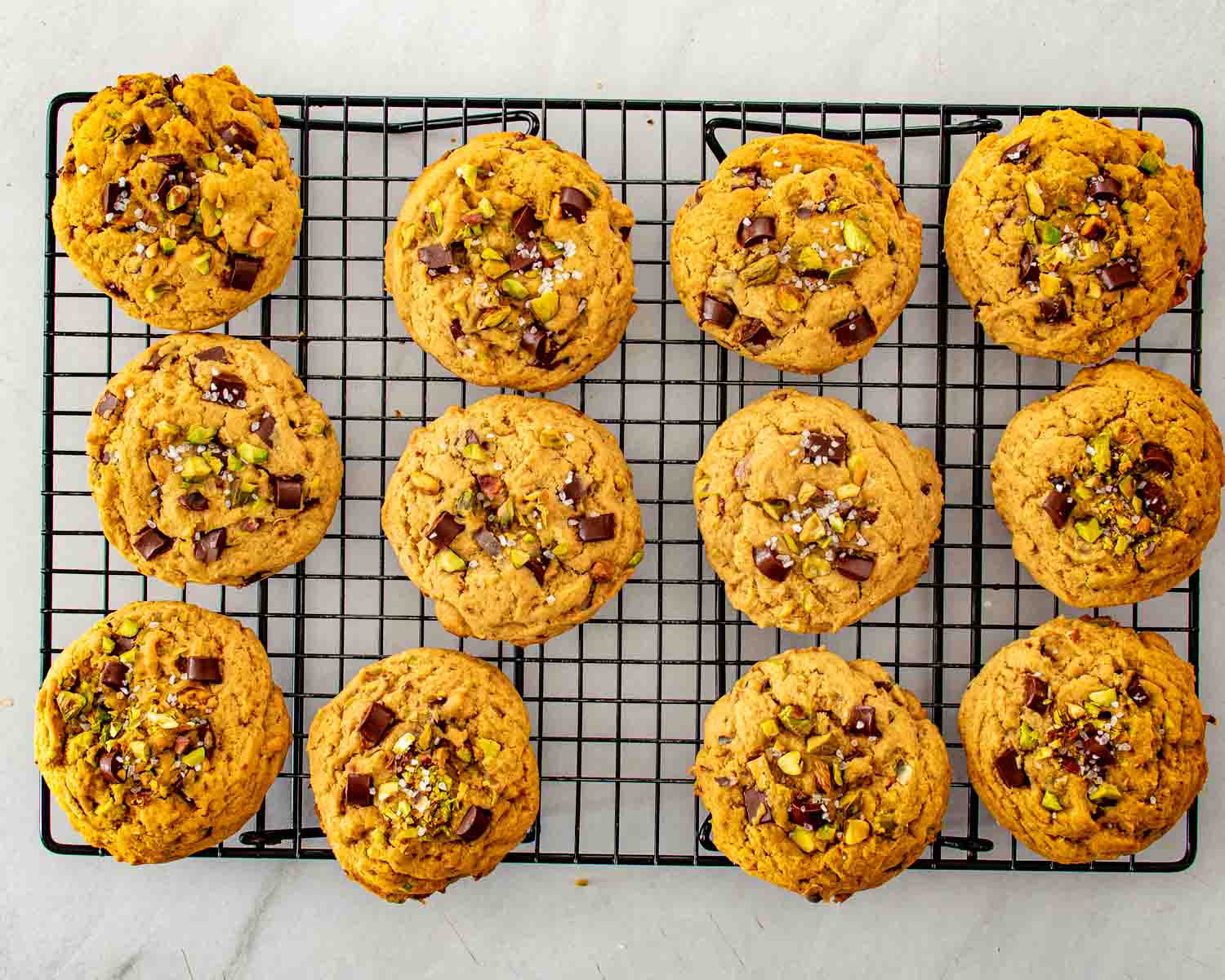 Freshly baked pistachio chocolate chunk cookies sprinkled with flaky sea salt cooling on a black wire rack.