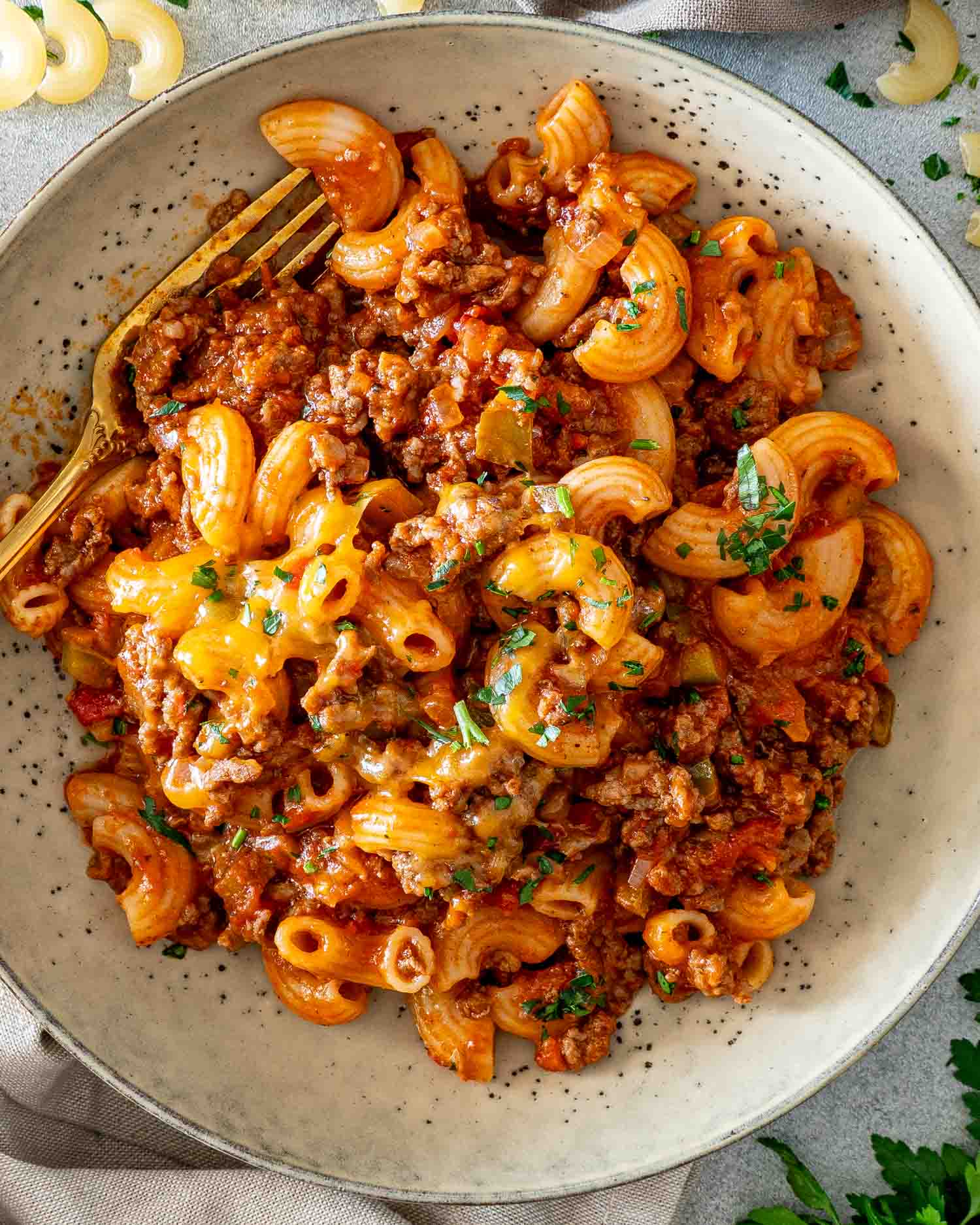 Overhead close-up of American goulash with elbow macaroni, ground beef, tomatoes, melted cheddar and parsley in a speckled bowl.
