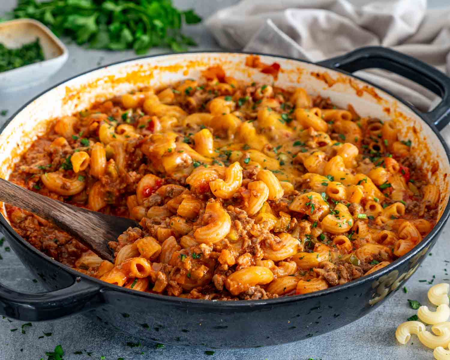 Wooden spoon holding beef and macaroni in a black enamel pot of American goulash topped with cheddar and parsley.