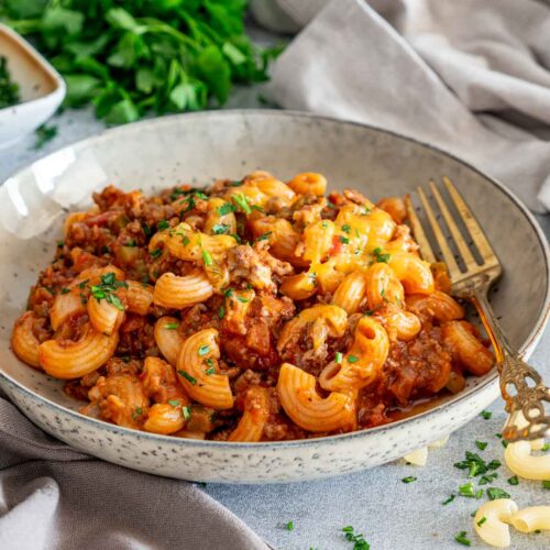 American goulash served in a shallow speckled bowl with an ornate gold fork, surrounded by parsley and uncooked macaroni.