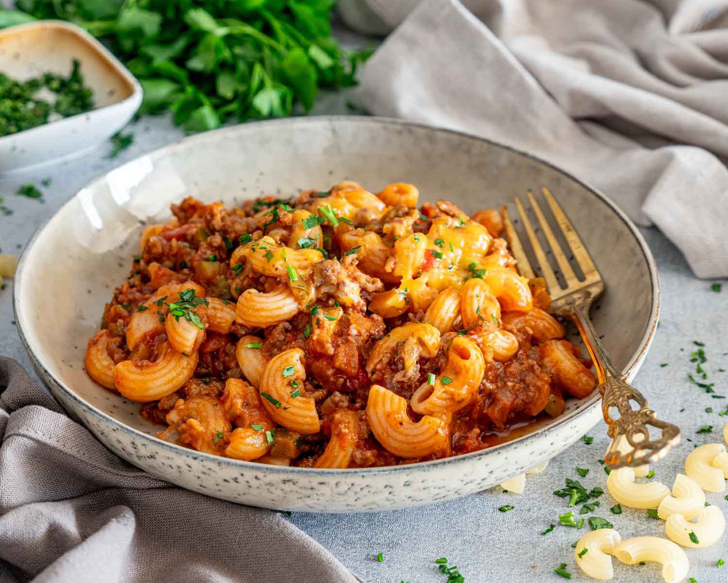 American goulash served in a shallow speckled bowl with an ornate gold fork, surrounded by parsley and uncooked macaroni.