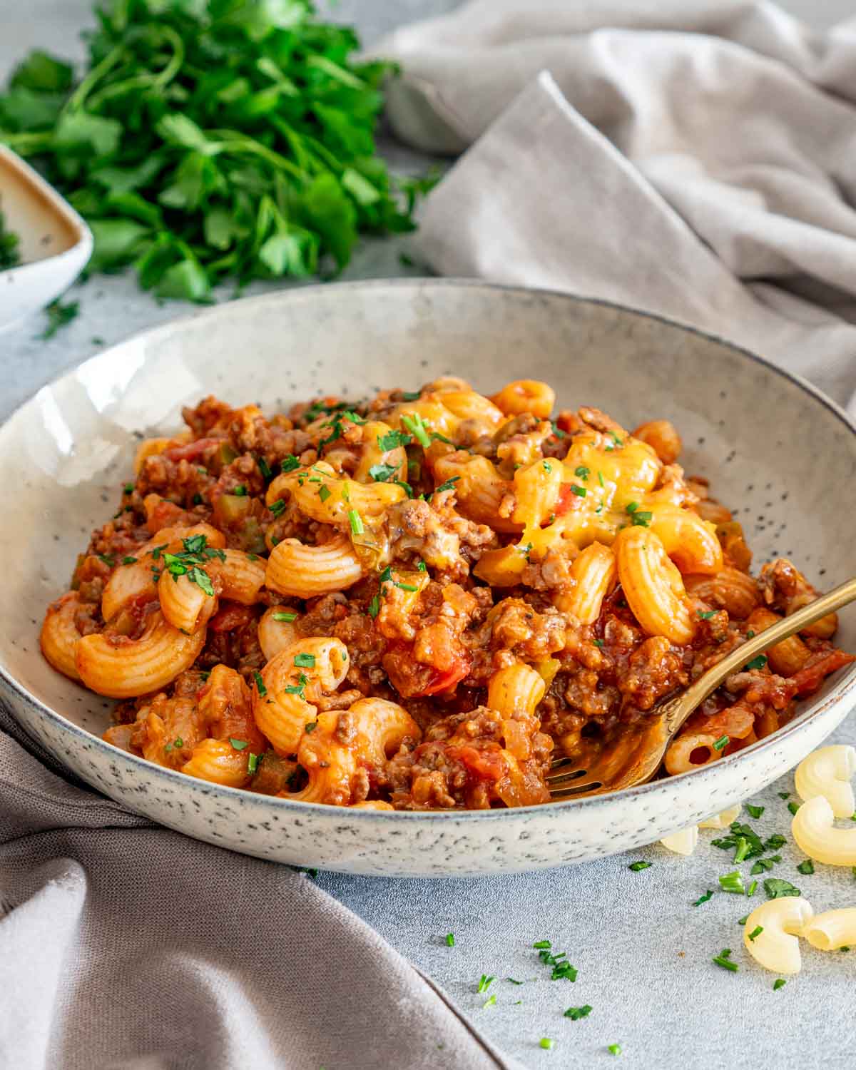 Close view of American goulash piled in a speckled bowl, topped with melted cheddar and parsley beside a gold fork.