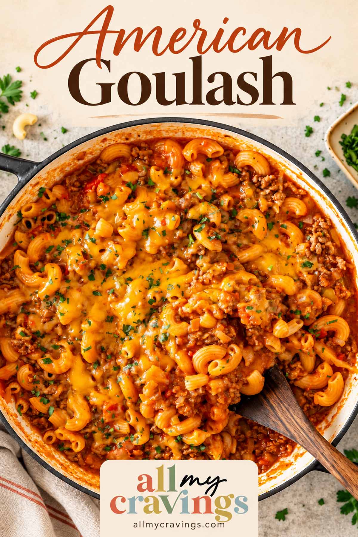 American Goulash title above a pot of cheesy tomato beef macaroni, with a wooden spoon and All My Cravings logo.
