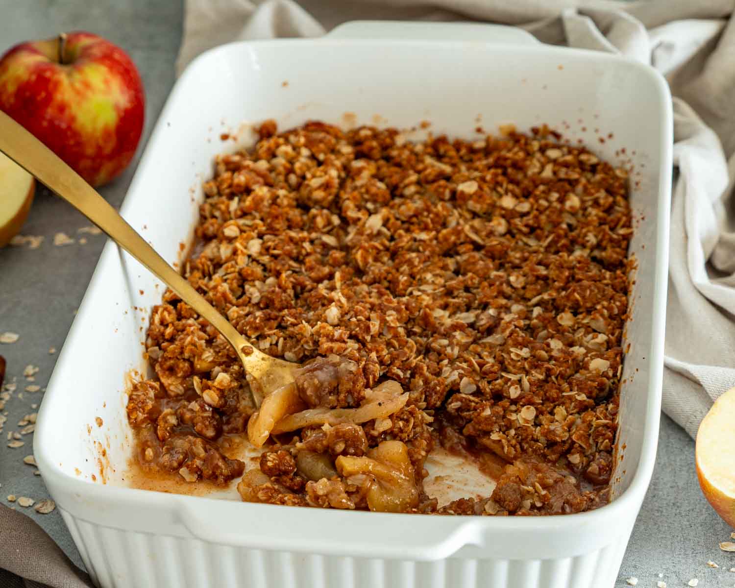 Golden apple crisp in a white baking dish with a spoonful removed to reveal tender cinnamon-spiced apples underneath.