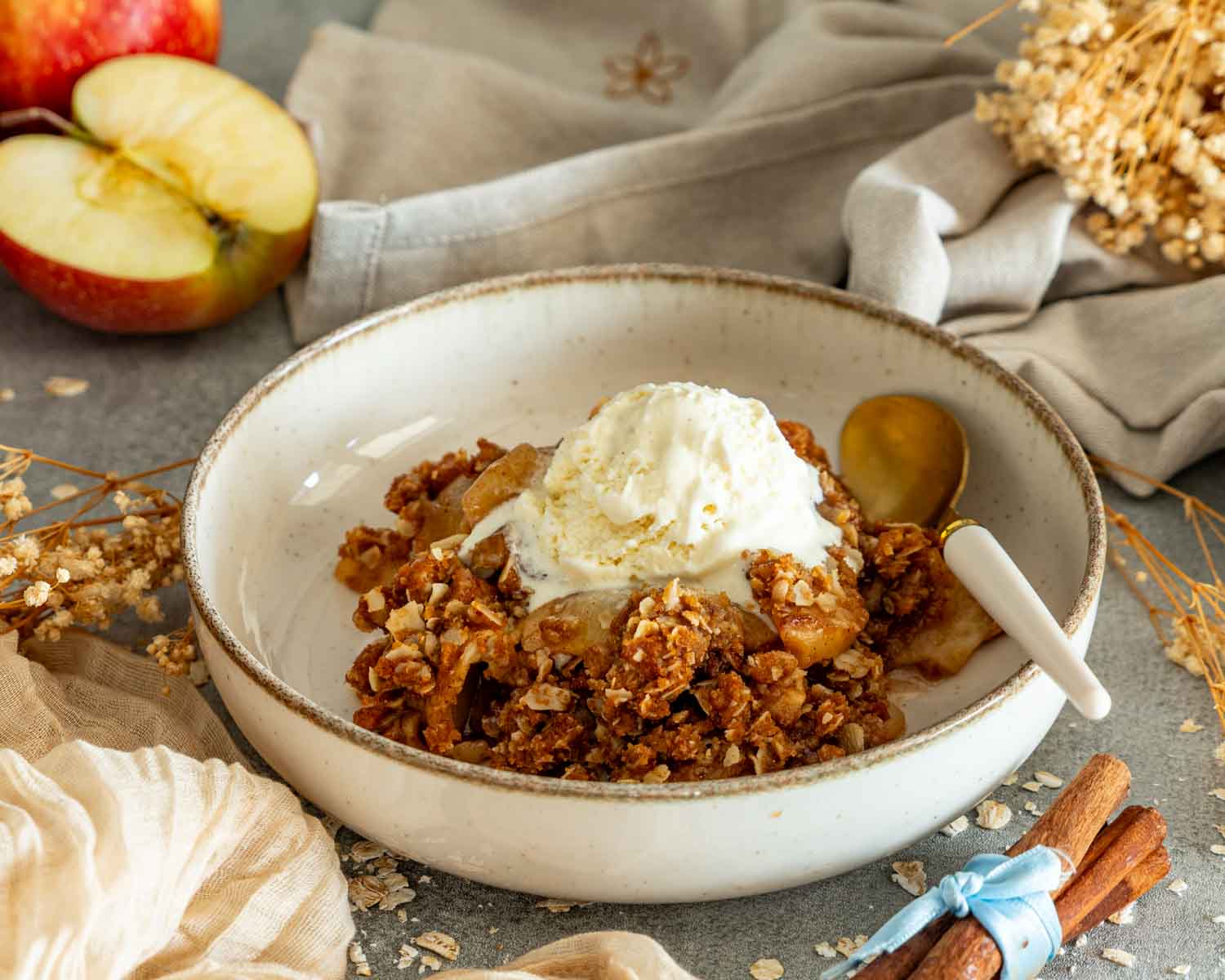 Bowl of homemade apple crisp topped with melting vanilla ice cream, styled with fresh apples, cinnamon sticks, and oats.