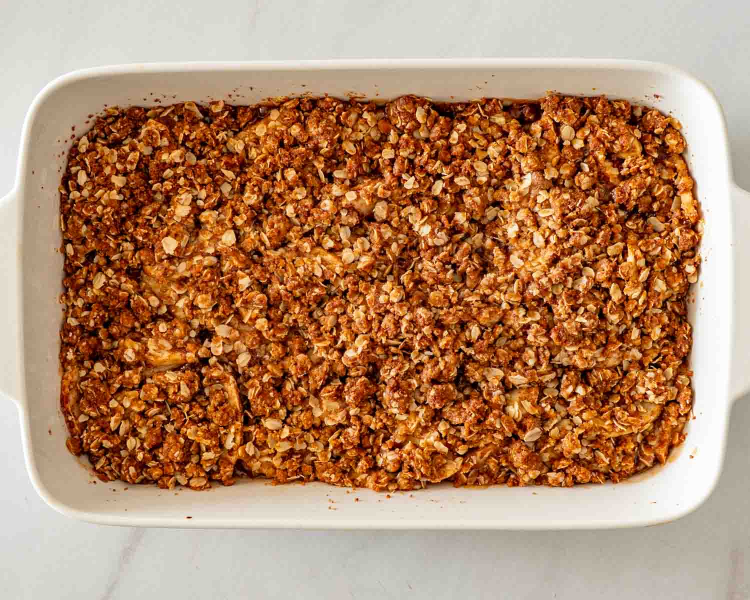 Overhead view of freshly baked apple crisp with a deep golden oat topping in a white rectangular baking dish.
