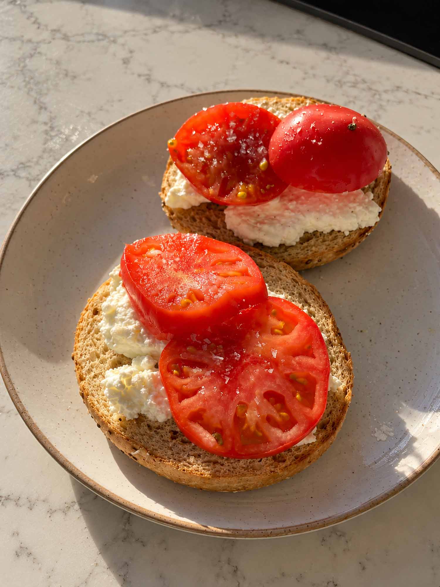 Toasted bagel halves topped with pressed cottage cheese, fresh tomato slices, and flaky Maldon salt on a ceramic plate.