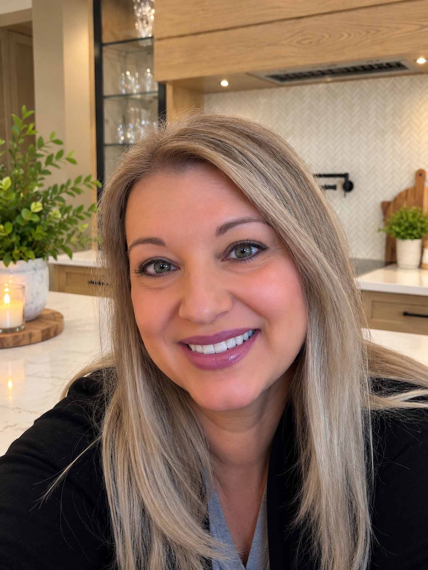 Joanna Cismaru smiling in her bright farmhouse kitchen, surrounded by white flowers, warm wood cabinets, and marble countertops.