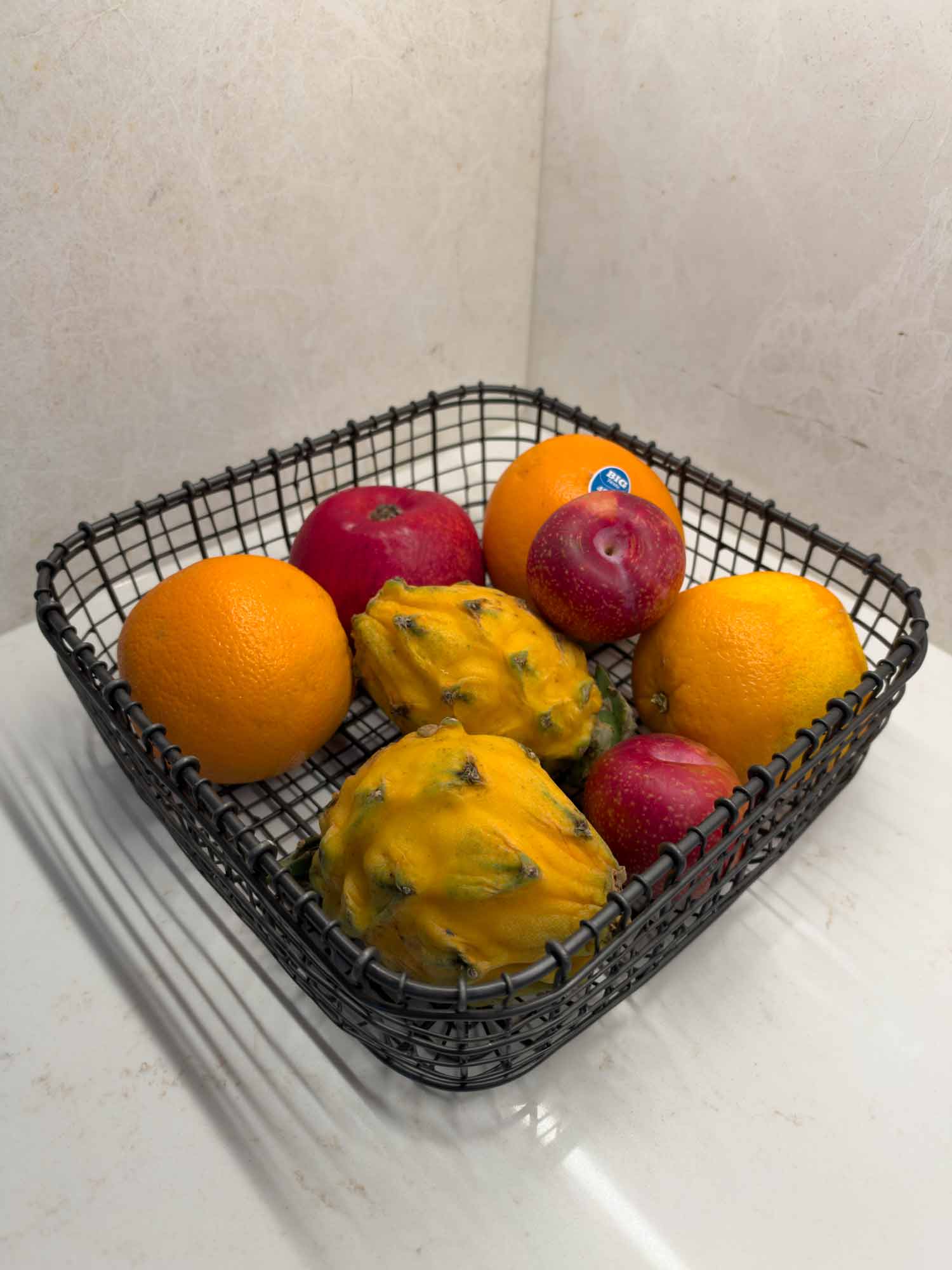 Black wire fruit basket filled with fresh oranges, red apples, and yellow dragon fruit on a white marble countertop.