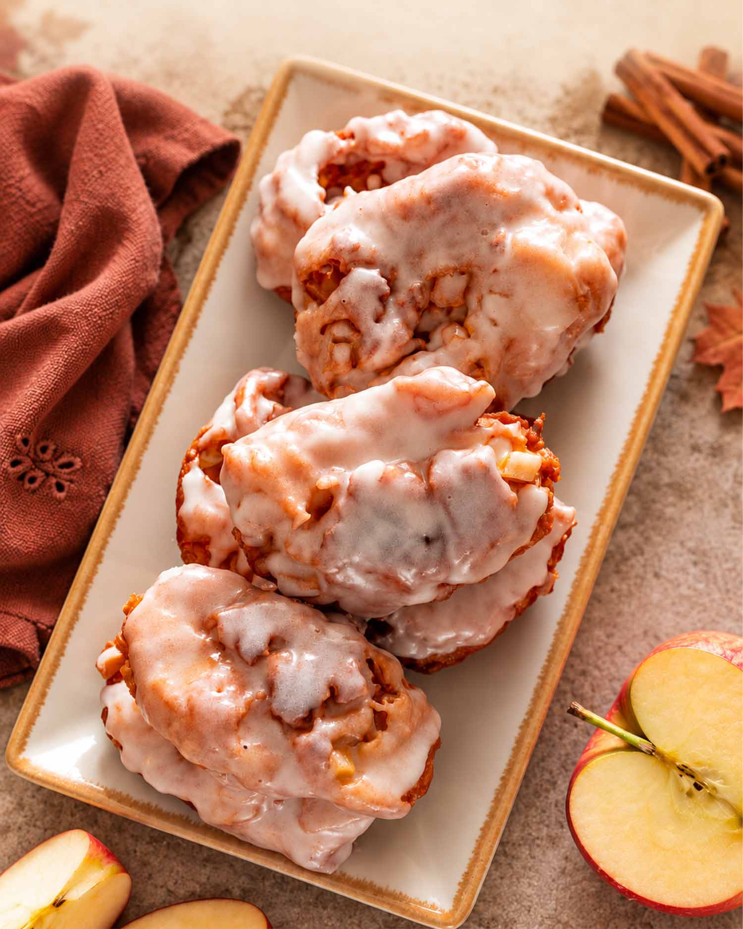 Glazed apple fritters arranged on a rectangular platter with sliced apples, cinnamon sticks, rust-colored linen, and autumn leaves.