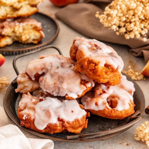 Stacked glazed apple fritters on a rustic metal tray, surrounded by sliced apples, cinnamon sticks, linen, and dried flowers.