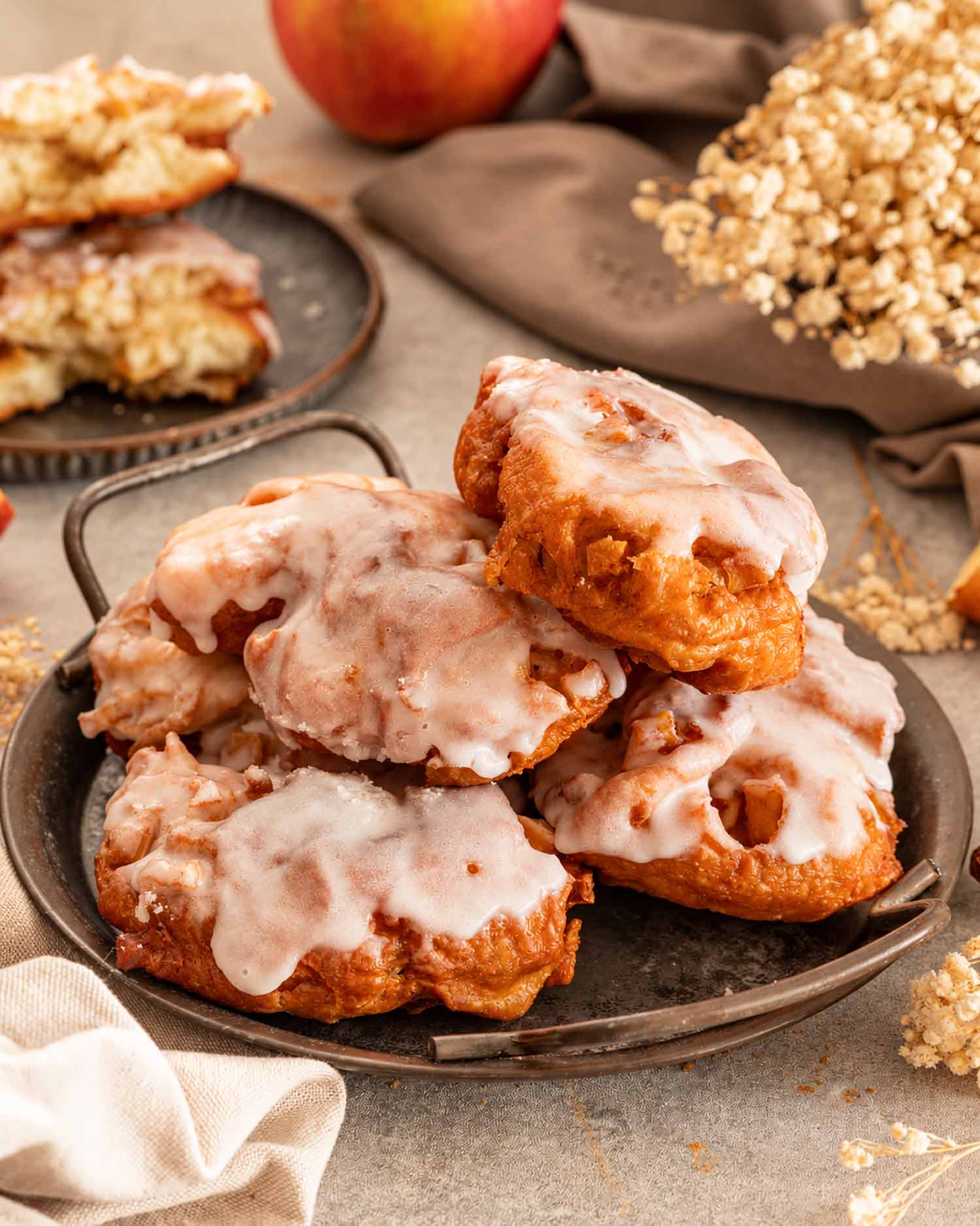 Close-up portrait of glazed apple fritters stacked on a metal tray with apples, linen, and dried autumn flowers.
