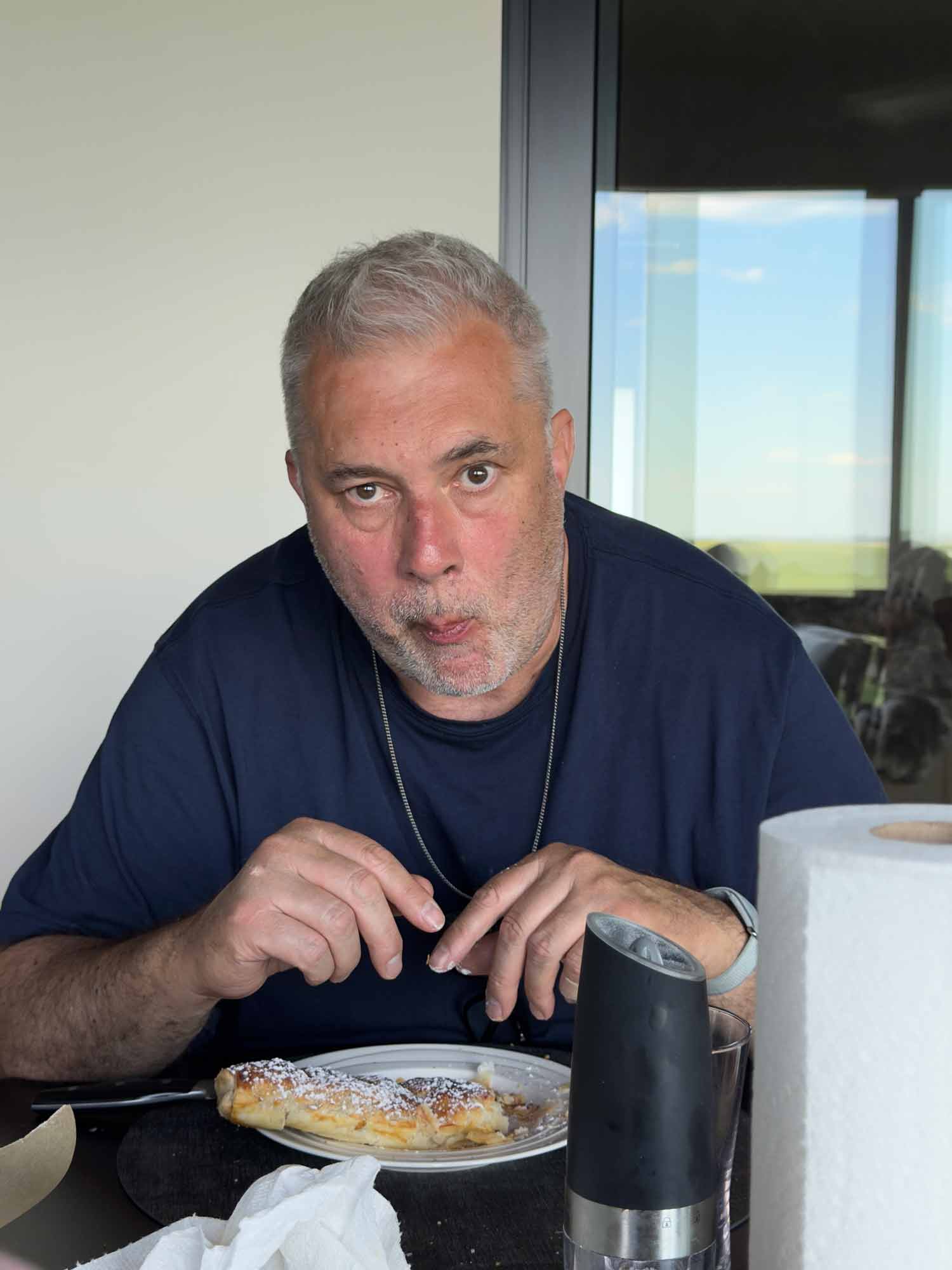 Jo&rsquo;s husband Remo sitting at the table, caught mid bite, eating a pastry dusted with powdered sugar.