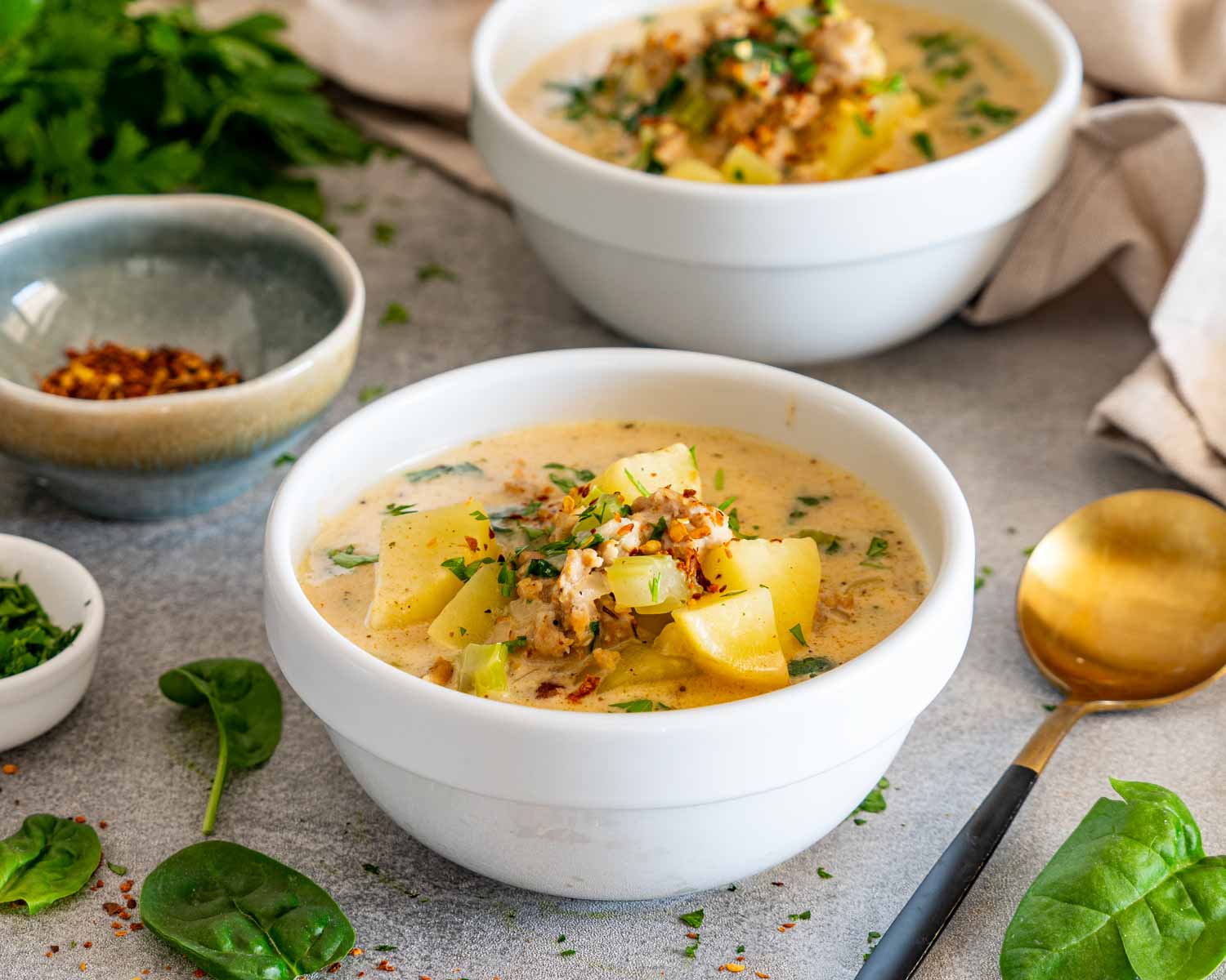 Bowl of creamy sausage potato soup topped with sausage, potatoes, parsley, and pepper flakes beside a gold spoon.