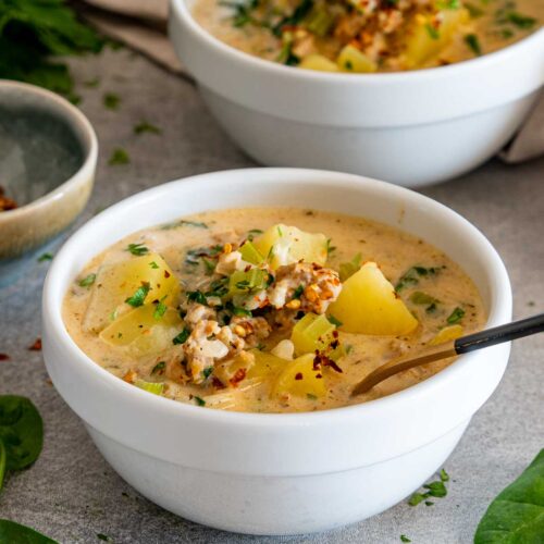 Close view of creamy sausage potato soup topped with tender potatoes, crumbled sausage, parsley, spinach, and red pepper flakes.