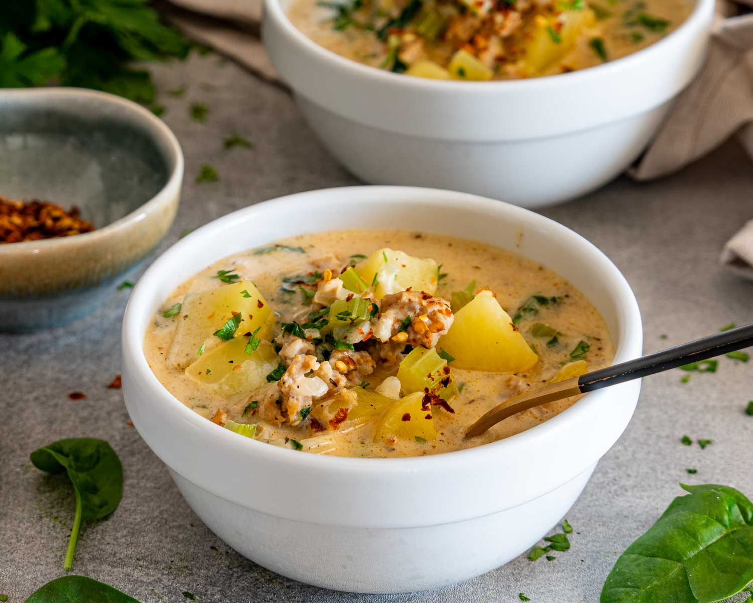 Close view of creamy sausage potato soup topped with tender potatoes, crumbled sausage, parsley, spinach, and red pepper flakes.