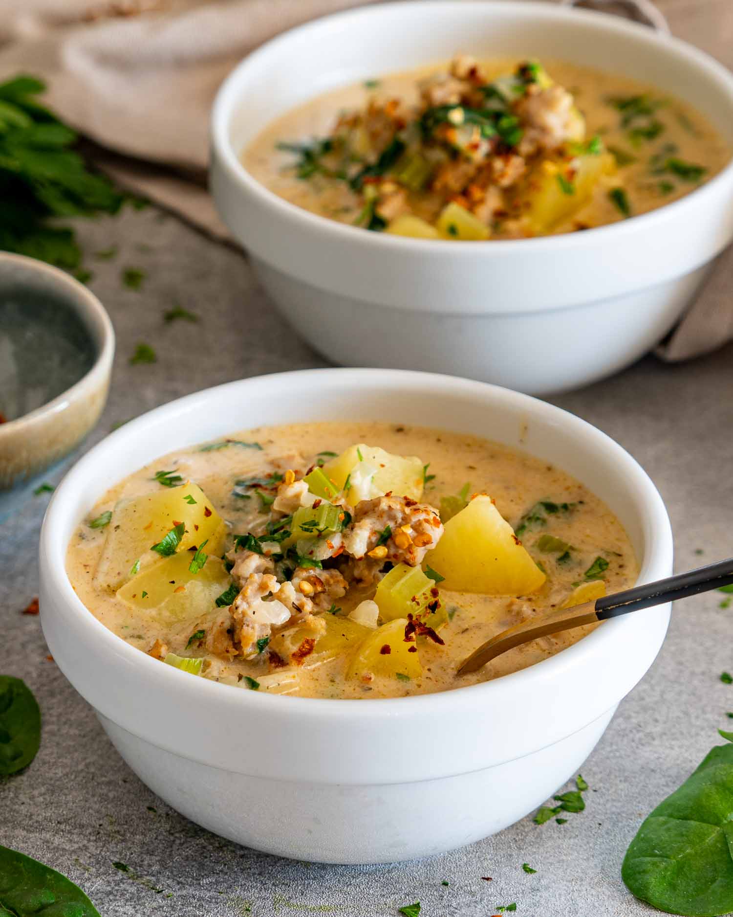Creamy sausage potato soup served in white bowls with tender potatoes, crumbled sausage, fresh herbs, spinach, and celery.