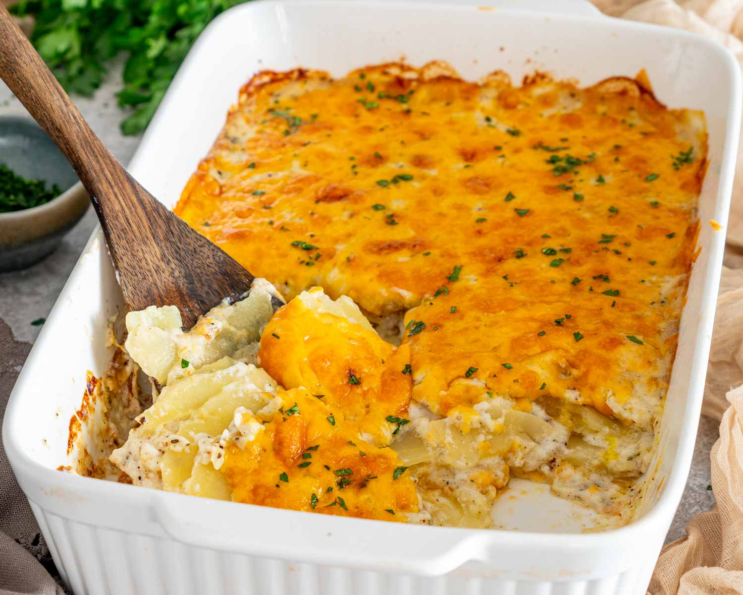 Scalloped potatoes being scooped from a white casserole dish, revealing tender potato layers beneath the golden cheddar topping.