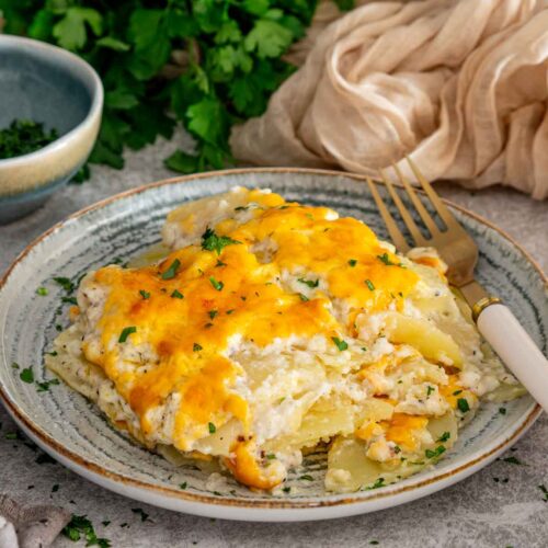 Serving of creamy, cheesy scalloped potatoes garnished with parsley and arranged on a plate beside a gold fork.