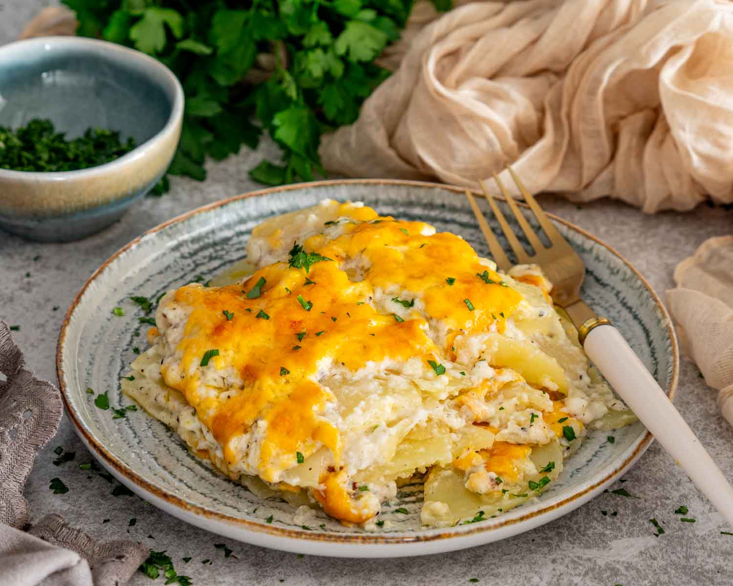 Serving of creamy, cheesy scalloped potatoes garnished with parsley and arranged on a plate beside a gold fork.