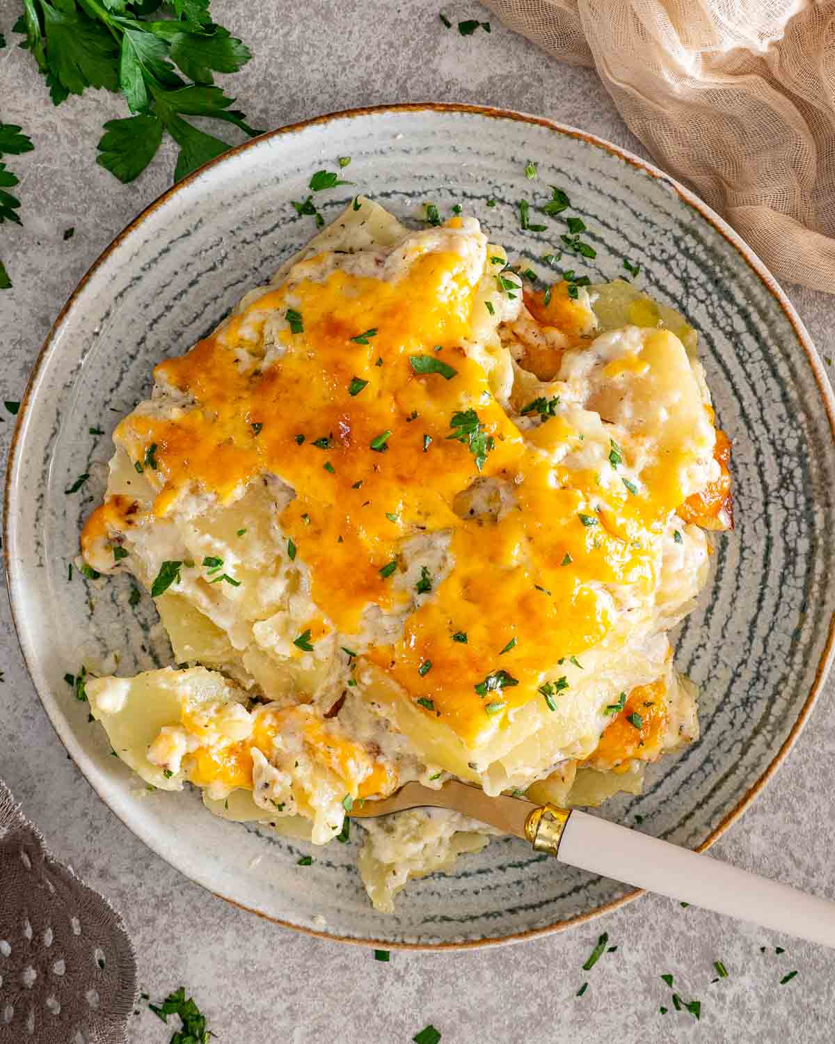Overhead view of creamy scalloped potatoes topped with melted cheddar and parsley on a rustic patterned plate.
