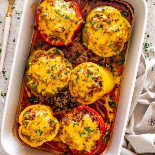 Six red, yellow, and green stuffed peppers topped with melted cheddar and parsley in a white baking dish.