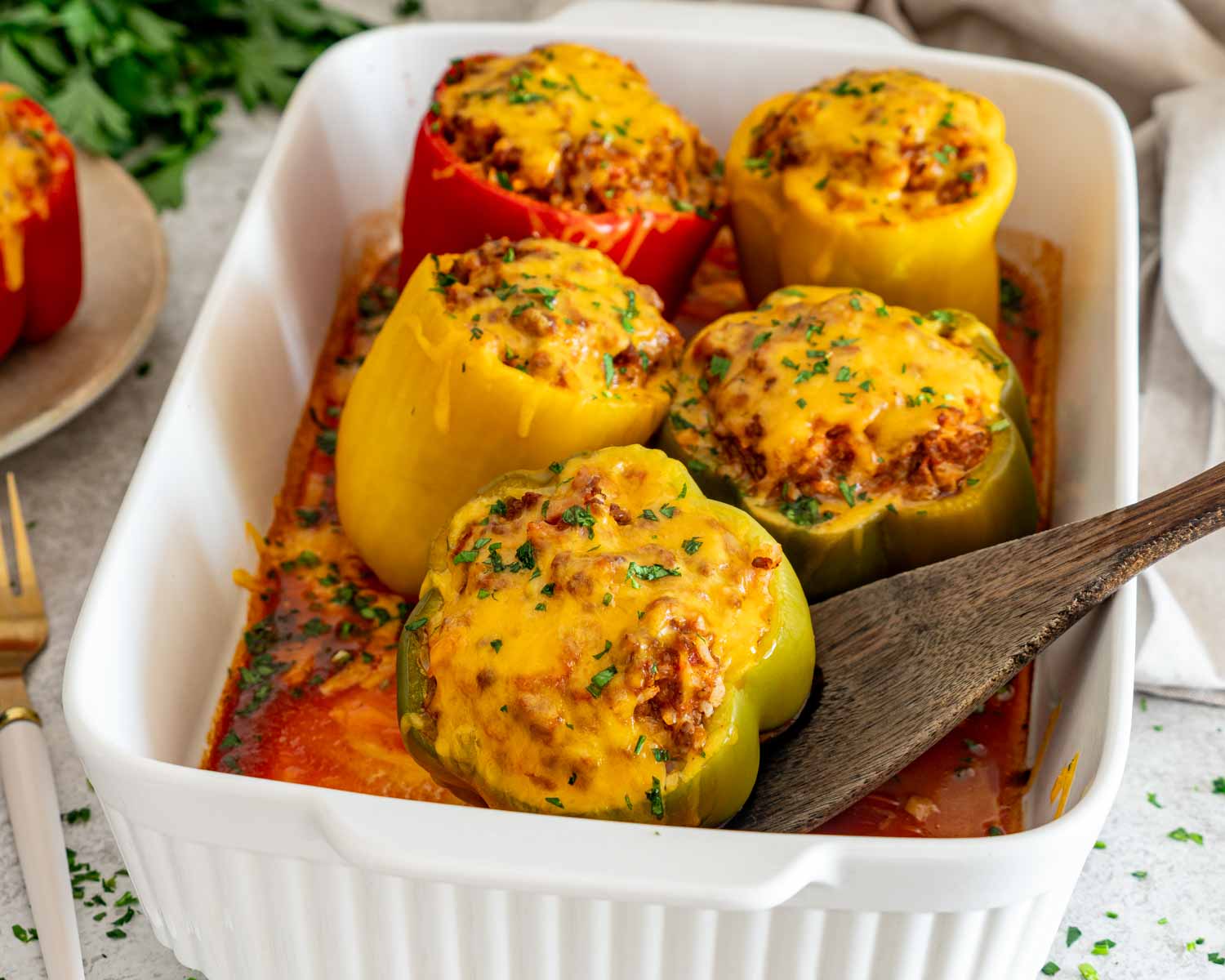 Cheesy baked stuffed peppers nestled in tomato sauce inside a white casserole dish with a wooden serving spoon.