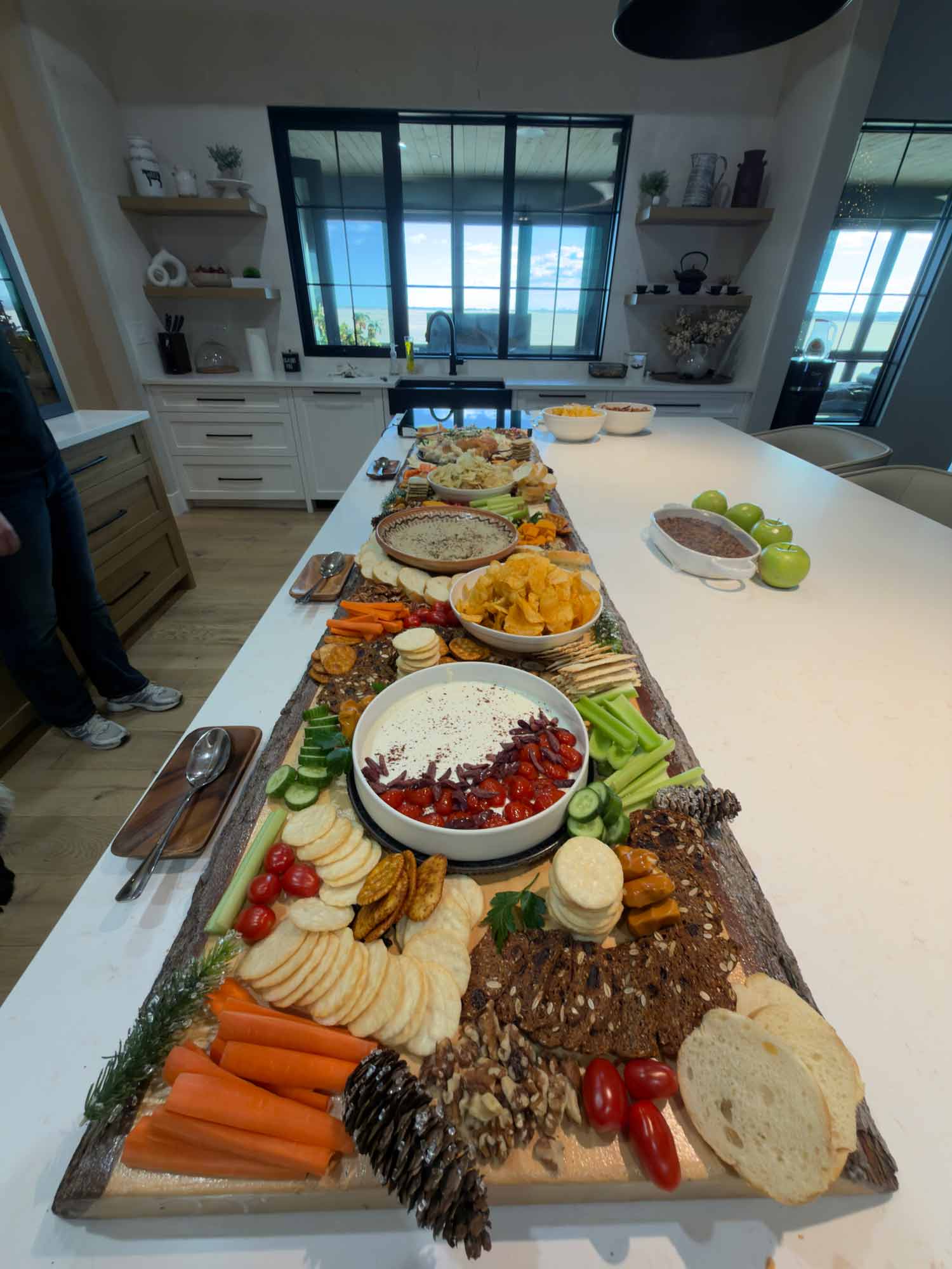 A long wooden grazing board on a kitchen island, filled with carrots, celery, cucumbers, cherry tomatoes, crackers, bread, walnuts, chips, and bowls of dip, set out for a Thursday get together.